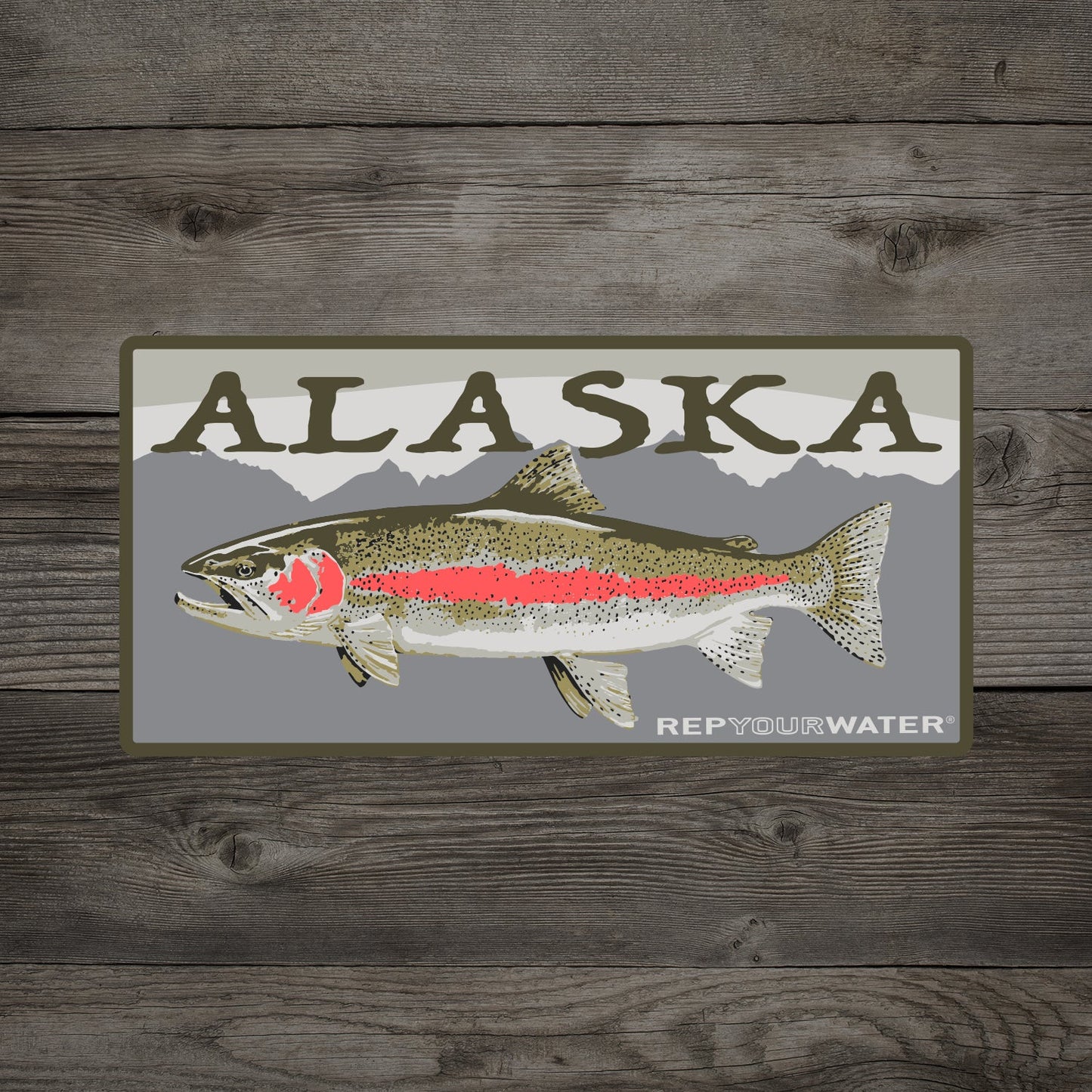 A wooden background with a mockup of a sticker is shown. The sticker says alaska with a rainbow trout on it