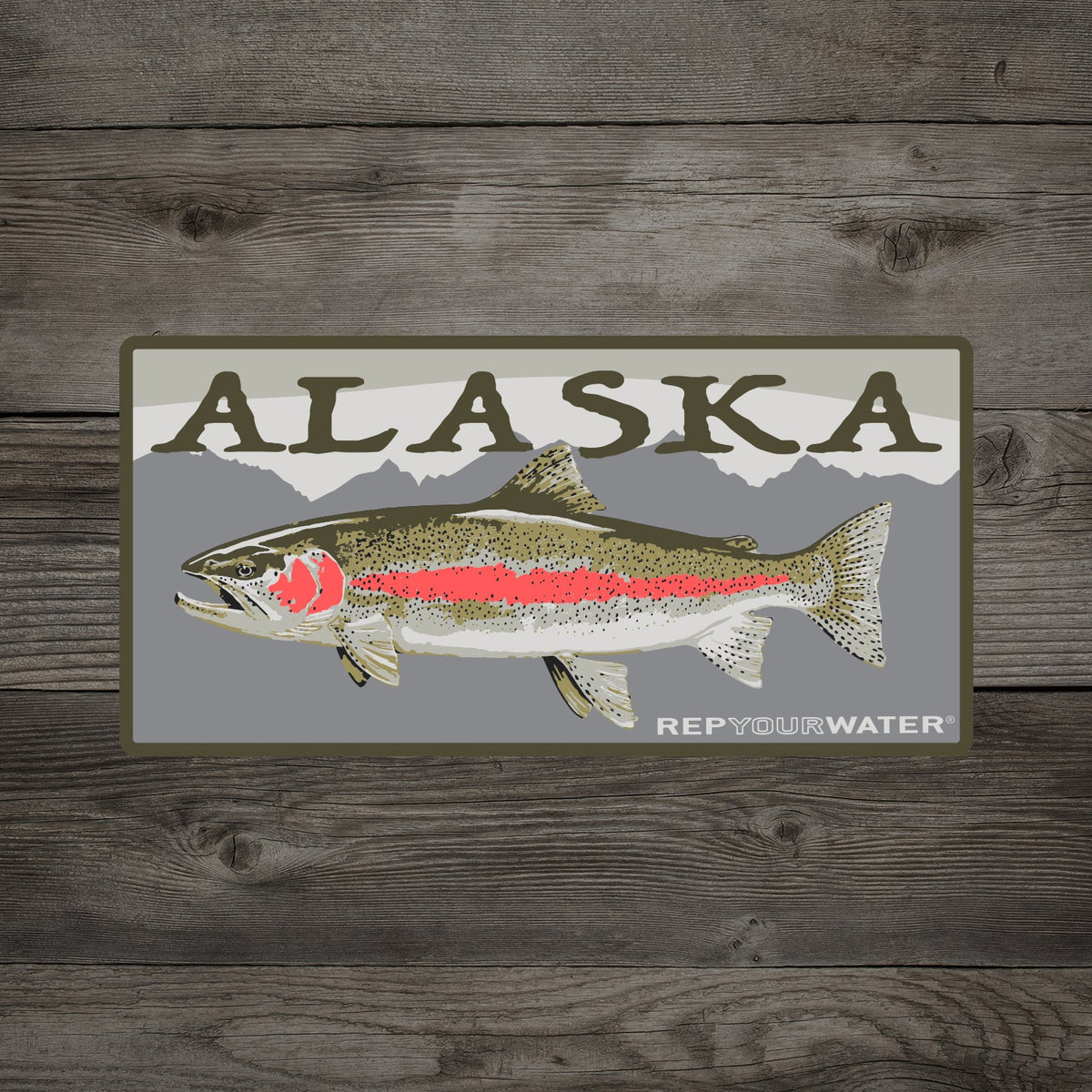 A wooden background with a mockup of a sticker is shown. The sticker says alaska with a rainbow trout on it