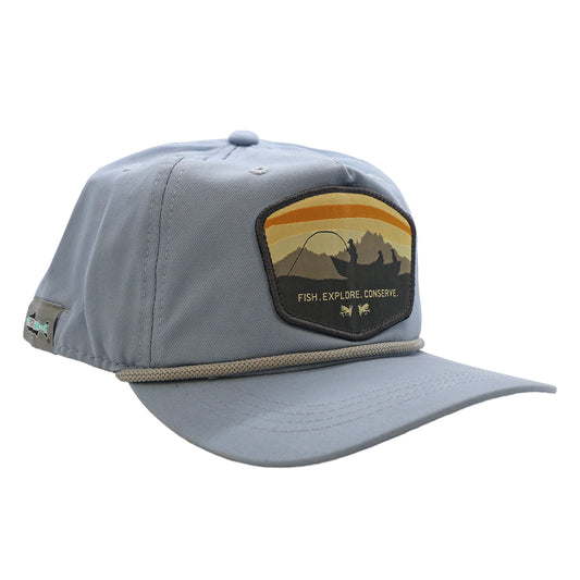 A unstructured 5 panel full cloth light blue hat. The front design says Fish Explore Conserve and has a boat with anglers and dog, 2 flies, and mountains with sunset skies