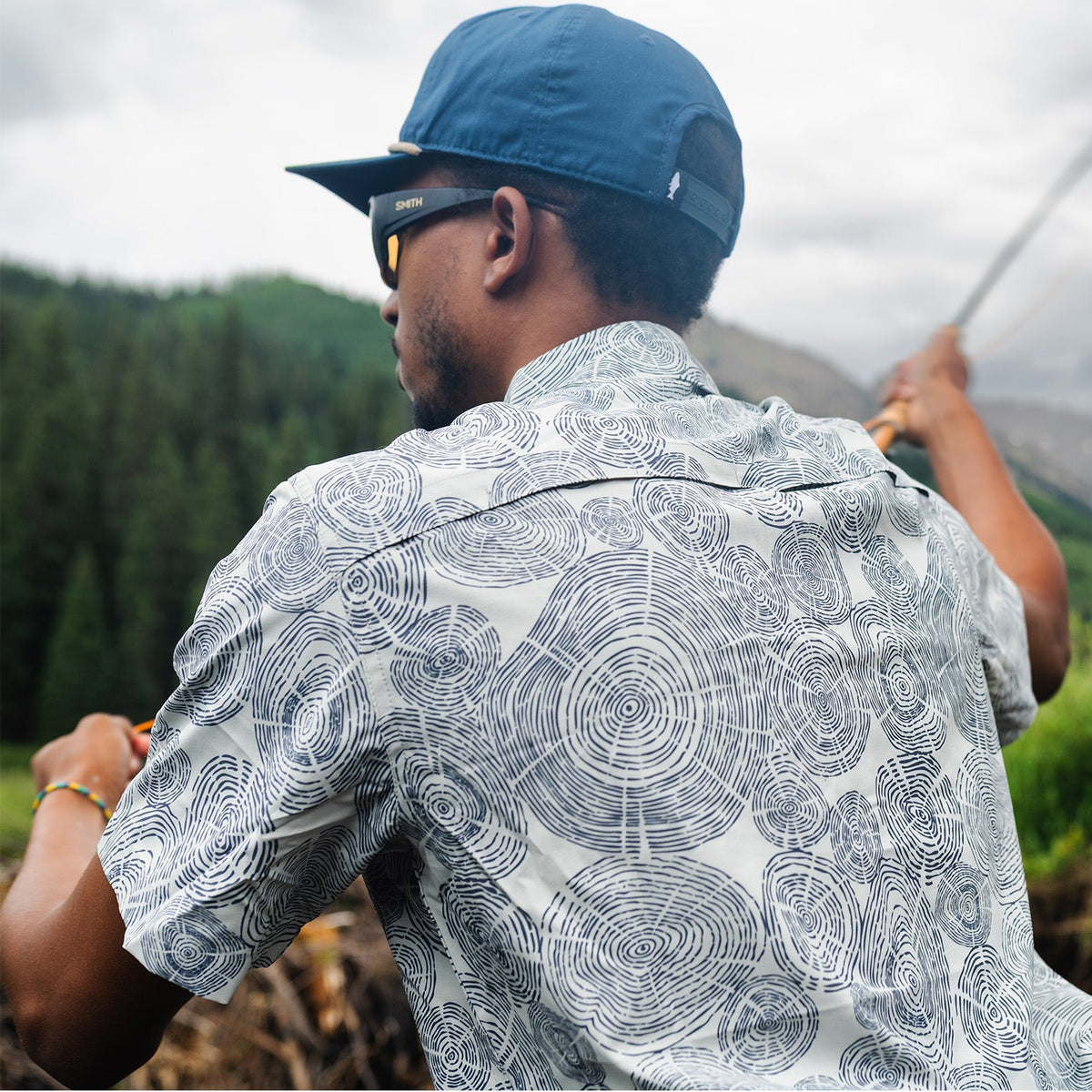 man casting wearing a blue hat and a gray shirt with tree ring pattern on it in the mountains