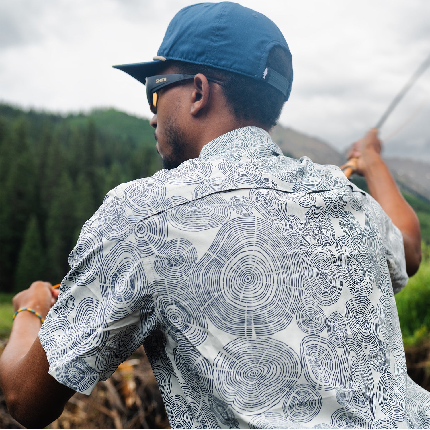 man casting wearing a blue hat and a gray shirt with tree ring pattern on it in the mountains