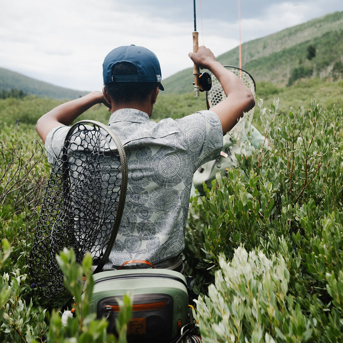 man walking through bushes wearing a blue hat and a gray deschutes shirt with tree ring pattern on it in the mountains