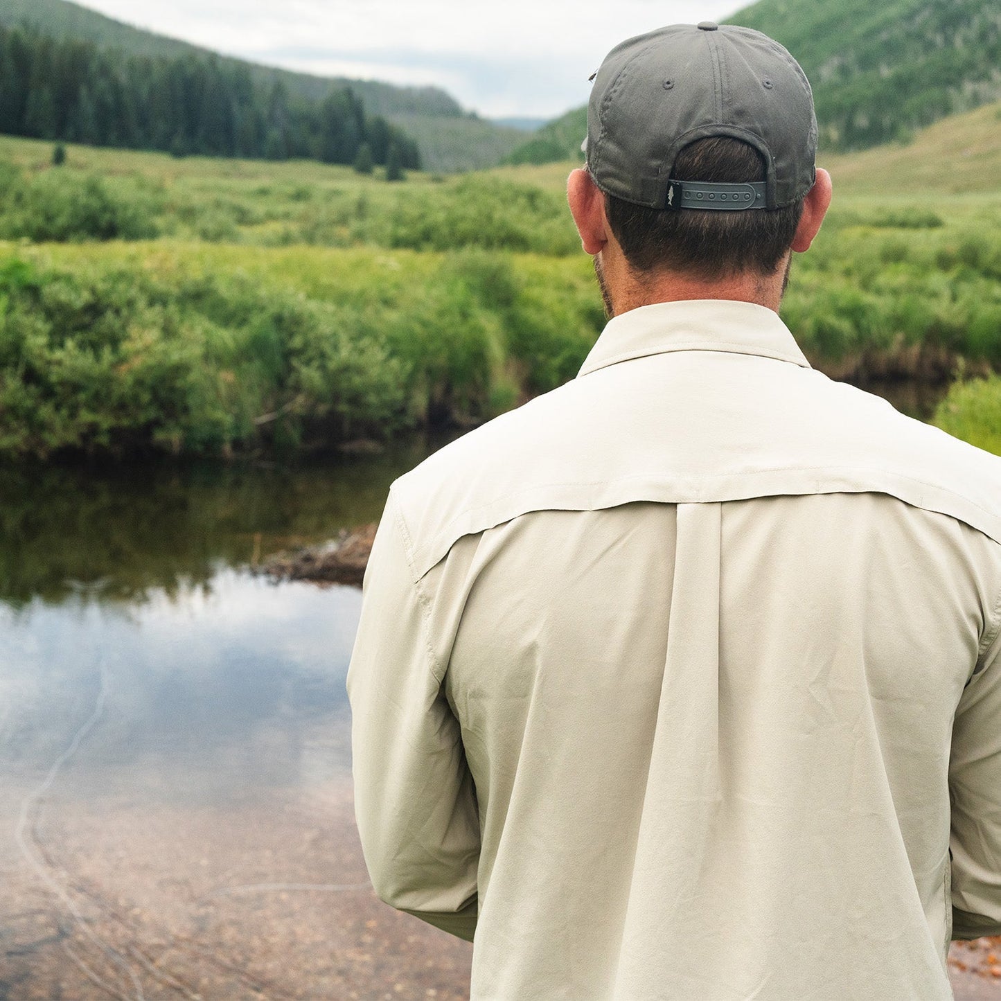 Man wearing fog gray button down with vented back standing by a mountain stream