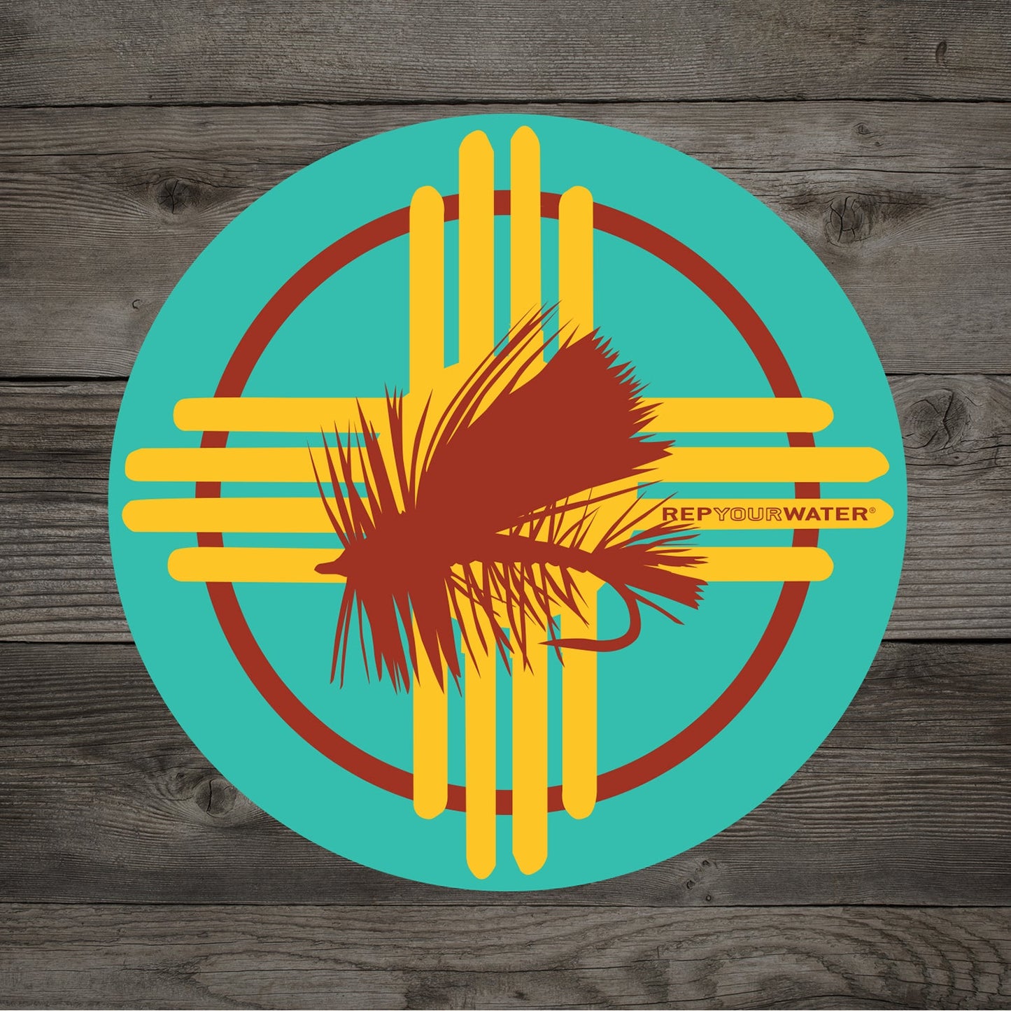 A wood background and circular teal sticker with the new mexico flag emblem and a red dry fly in the middle with text that reads rep your water